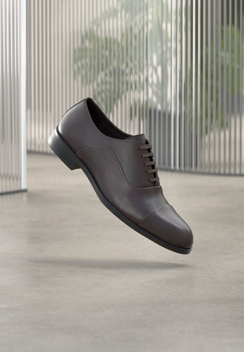 Barna bőrből készült elegáns cipő kerek orral, fűzős kialakítással, sima felülettel és lapos fekete talppal, semleges háttér előtt lógva.