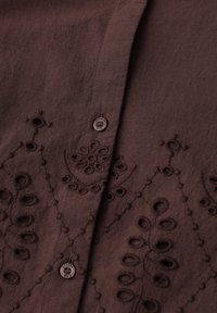 Tissu marron avec broderie florale découpée et détails cousus, comprenant deux boutons ronds le long d'une couture centrale.