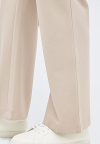 Μπεζ παντελόνι με φαρδιά μπατζάκια συνδυασμένο με λευκά δερμάτινα αθλητικά παπούτσια σε φωτεινό φόντο, εμφανίζοντας τα κάτω πόδια και τα πόδια.