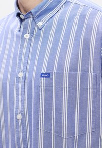 Hellblau gestreiftes Button-up-Hemd mit weißen vertikalen Streifen. Verfügt über eine Brusttasche mit einem blauen Etikett mit der Aufschrift "Hugo." Baumwollstoff, mit Kragen.
