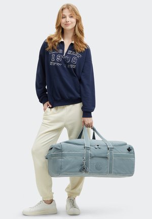 Blue duffel bag with dual handles, stitched accents, and a decorative keychain. Model wears a navy hoodie and cream joggers with white shoes.