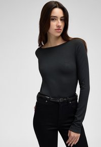 Naine, kes kannab keha rõhkuvat musta soonikkoega pika varrukaga pluusi ja musti pükse tekstuurse vööga, seistes ühevärvilise helehall tausta ees.