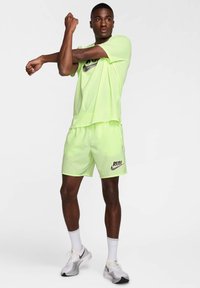 Conjunto deportivo de color verde neón que incluye una camiseta holgada y pantalones cortos. Presenta el logotipo negro "RUN" en los pantalones cortos, combinados con zapatillas blancas y grises.