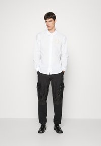 Camicia bianca a maniche lunghe con bottoni e un emblema rotondo, abbinata a pantaloni cargo neri con tasche con cerniera e una texture leggermente lucida.