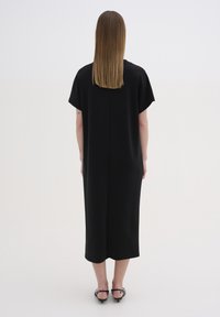 Černé oversized šaty s krátkým rukávem, rovný střih, délka do poloviny lýtek, hladká textura, středový šev na zadní straně a rozparek na spodním okraji.