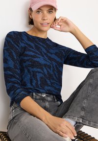 Frau trägt eine rosa Mütze, ein langärmliges Hemd mit blauem und schwarzem Muster und graue Jeans. Sie sitzt mit einem Arm, der auf ihrem Knie ruht, an einer weißen Wand.