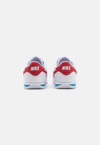 Nike sportbačiai, turintys baltą viršutinę dalį, raudonus akcentus, mėlyną detalę ant kulno ir išsiskiriantį "NIKE" logotipą ant kulno lipduko.