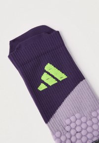 Violeta un lavandas sporta zeķe ar reljefiem saķeres punktiem un zaļu Adidas logotipu pie aproces, attēlota uz balta fona.