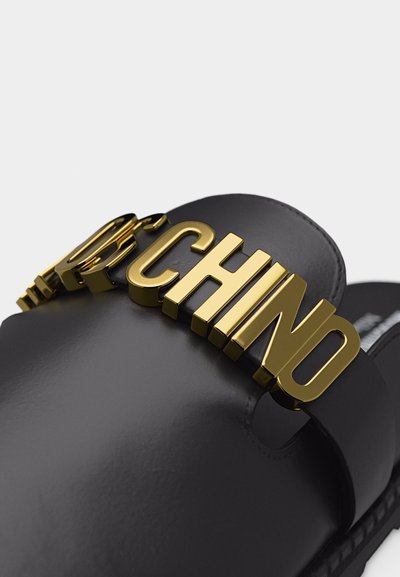 Μαύρο δερμάτινο σανδάλι τύπου slide με μεγάλα χρυσά μεταλλικά γράμματα που σχηματίζουν τη λέξη «MOSCHINO» στον ιμάντα.