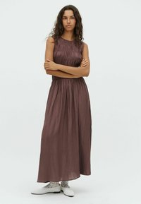 Maxiklänning i mörk mauve, tillverkad av textil med strukturerad yta och rynkdetaljer, ärmlös design, åtsittande midja, flödande kjol, bärs med vita skor.