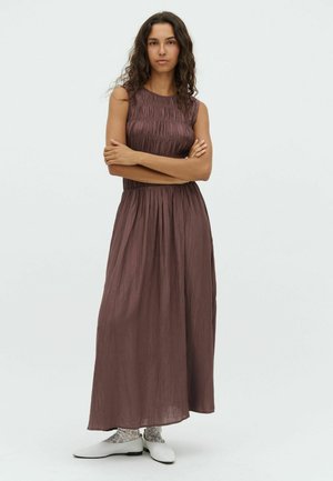 Maxi-Kleid in dunklem Mauve, aus strukturiertem Stoff mit gerafften Akzenten, ärmelloses Design, tailliert, fließender Rock, getragen mit weißen Schuhen.