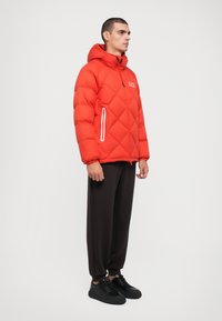 Red puffer jacket with diamond quilted design, a hood, and white zippered pockets. Paired with black loose-fit pants and dark sneakers.