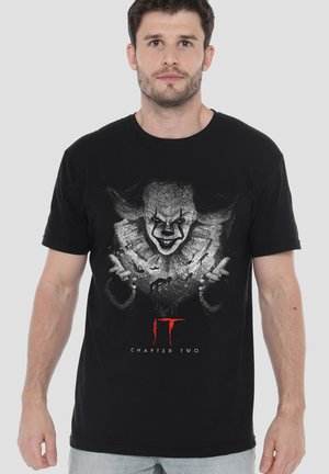 Man wearing black T-shirt featuring menacing clown face, extended hands, and "IT Chapter Two" text on front, standing against plain background.