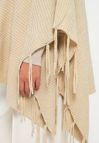Poncho bege tricotado com nervuras texturizadas e bordas franjas. Apresenta um design drapeado, com detalhes de costura intrincados.