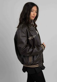 Veste aviateur en cuir brun avec des poignets et un ourlet côtelés. Présente des broderies dorées sur la poitrine gauche et des poches latérales. Texture lisse, coupe oversize.