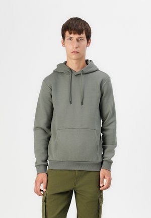 Olive green cotton hoodie with a front pocket, drawstring hood, and ribbed cuffs. Worn with cargo pants. Simple, minimalist design.