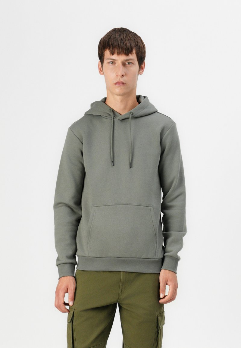 Olive green cotton hoodie with a front pocket, drawstring hood, and ribbed cuffs. Worn with cargo pants. Simple, minimalist design.