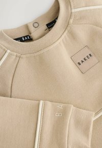 Beżowa bluza z okrągłym kołnierzem, białymi detalami szwów, zapięciem na zatrzask przy szyi oraz małą naszywką "BAKER" na piersi.