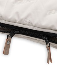 Close-up van een beige stof met kleine reliëfpatronen en een zwarte dubbele rits met bruine leren trekknoppen.