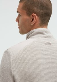 Hellgrauer Pullover mit strukturiertem Strickmuster und glatten Schulterpaneelen. Hoher Kragen und Markenlabel auf der Rückseite. Lässige Passform.