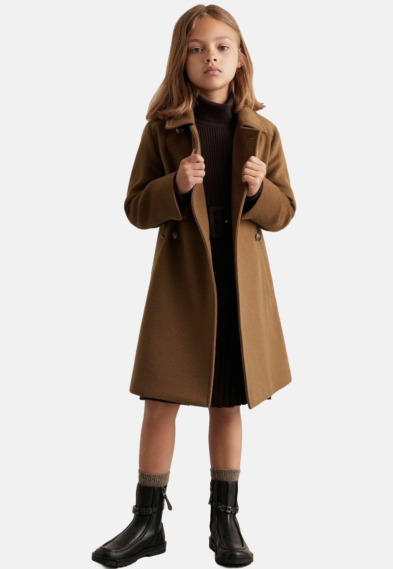 Rudas vilnonis paltas su pritaikytu dizainu, dvigubais mygtukais ir dviem šoninėmis kišenėmis. Dėvimas ant tamsios raukšlėtos suknelės ir juodų botų.