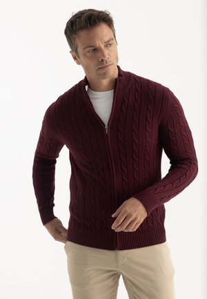 Homme ajustant un pull zippé bordeaux en maille torsadée, portant une chemise blanche et un pantalon beige, debout devant un fond clair uni.