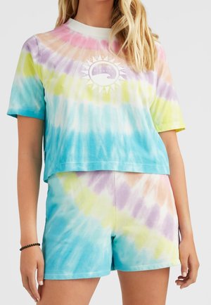 Kobieta ubrana w pastelowy krótki top z krótkim rękawem w stylu tie-dye oraz dopasowane szorty z logo słońca i fali na piersi.