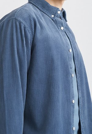 Osoba ubrana w niebieską koszulę sztruksową na guziki, na jasnoniebieskiej bluzce, ukazująca fakturę i szczegóły materiału koszuli oraz guzików.