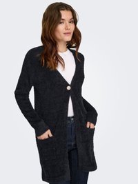 Pruunide juustega naine, kes kannab musta pehmet kardigani, millel on üks dekoratiivne nööp, valget särki ja tumesiniseid teksaseid, käed kardigani taskutes.