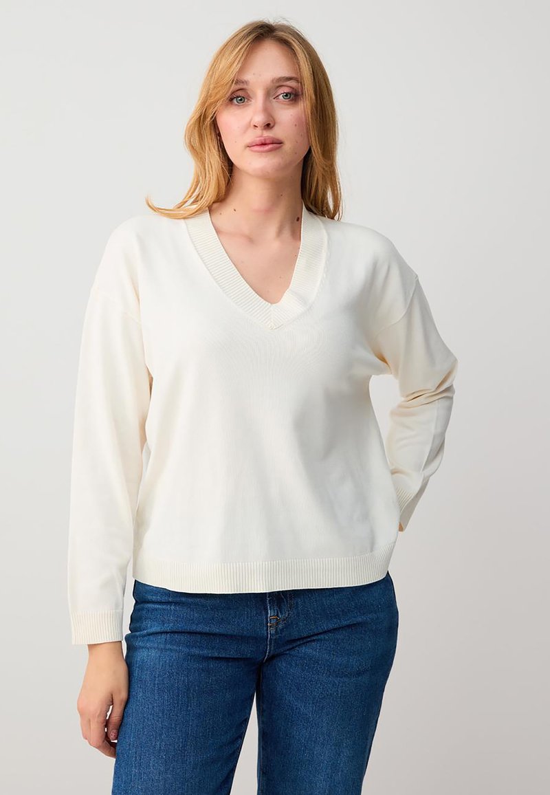 Femme blonde debout, la main sur la hanche, portant un pull blanc à col en V et un jean bleu, sur un fond neutre.