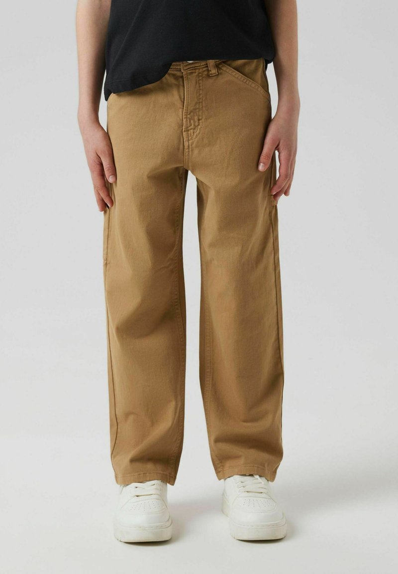 Pantalones casuales color beige, confeccionados en algodón, con un corte relajado, bolsillos laterales y cierre de cremallera, combinados con zapatillas blancas y una blusa negra.