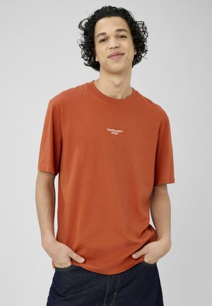 Jeune homme aux cheveux bouclés portant un T-shirt couleur rouille avec le texte "TEMPÉRAMENT DE FEU", les mains dans les poches d'un jean bleu foncé, fond neutre.