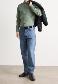 Groene button-up shirt, blauwe denim jeans, zwarte riem en zwarte leren laarzen. Zwarte jas over schouder gedrapeerd, staand op een lichte vloer.