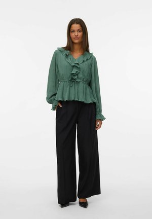 Blouse verte plissée avec un col et des poignets volantés; taille cintrée, associée à un pantalon noir à jambes larges.