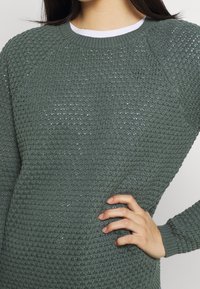 Grüner gestrickter Pullover mit einem strukturierten Muster, Raglanärmeln und einem kontrastierenden weißen Ausschnitt. Weiche, gerippte Bündchen runden das Design ab.