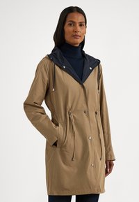 Světle hnědá lehká bunda s tmavě modrou kapucí, šňůrkami, patentními knoflíky a bočními kapsami. Hladká textura, design sahající až ke kolenům pro ochranu před počasím.