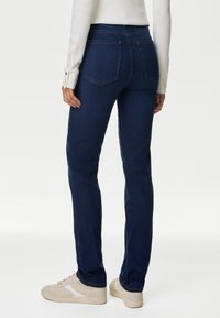 Jeans de mezclilla azul oscuro con una pierna ligeramente ajustada, diseño de cinco bolsillos y detalles de costura mínimos. Combinados con zapatillas de color claro.