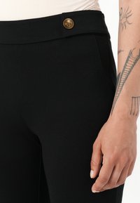 Pantaloni neri a vita alta realizzati in tessuto testurizzato, con dettaglio in bottone metallico e una finitura liscia. Nessuna tasca visibile.