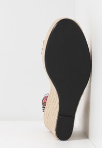 Vue de dessous d'une sandale espadrille compensée avec semelle noire et semelle intermédiaire en jute tissée sur fond blanc.