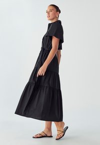 Abito maxi nero con un design a strati, maniche corte e vita con cintura. Il tessuto è liscio con una leggera lucentezza. Accompagnato da sandali neri minimal.