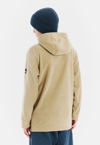 Beige fleece hoodie met een grote capuchon, geribde marineblauwe beanie en een gestructureerd oppervlak. Voorzien van een zwart logo op de linker mouw.