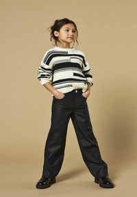 Maglione a righe lavorato a maglia in bianco e nero, abbinato a pantaloni neri a gamba larga e scarpe nere lucide. Il modello sta in piedi con le mani nelle tasche.