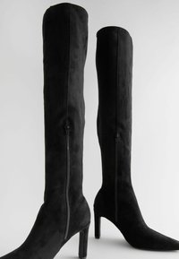 Schwarze kniehohe Stiefel aus weichem Wildleder, mit einem eleganten Design, spitzem Zehenbereich und einem Seitenreißverschluss. Der Stiletto-Absatz sorgt für Höhe.