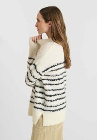 Maglione di grandi dimensioni di colore crema con righe orizzontali nere, dotato di un collo alto e spacchi laterali. Tessuto adatto alla maglia morbido e testurizzato.
