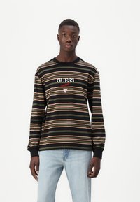 Svart langermet skjorte med horisontale striper i krem, rosa og oliven. Midt på brystet står logoen "GUESS originals" i hvitt og rødt.