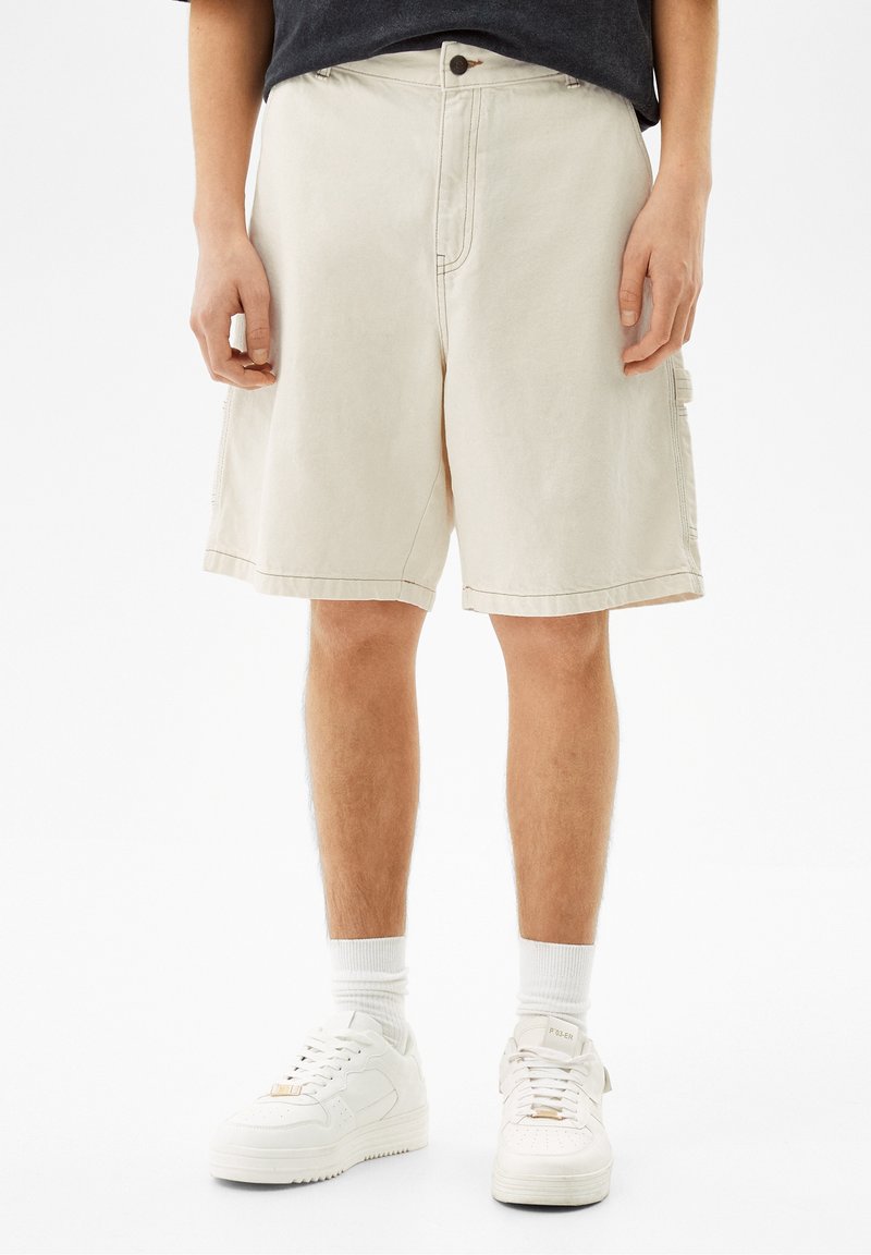 Personne portant un short beige jusqu'aux genoux, des chaussettes blanches montantes et des baskets blanches, debout devant un arrière-plan uni.