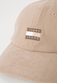 Beige corduroy-baseball-lippalakki, jossa on suorakulmainen tarra, jossa on teksti "TOMMY JEANS" ja vaakasuorat väriraidat valkoisena ja vaaleanruskeana.