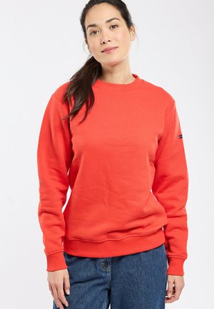 Femme aux cheveux bruns attachés en queue de cheval, portant un sweat-shirt à manches longues rouge vif et un jean bleu, debout devant un fond blanc.