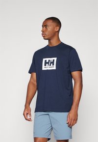 Tumed sinine puuvillane T-särk, millel on valge trükitud logo, mis ütleb "HELly HANSEN" ristkülikukujulises disainis. Lühikesed varrukad, ümar kaelus.