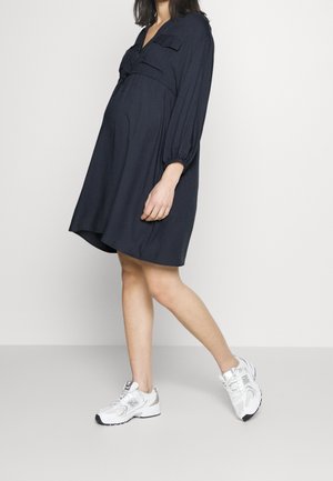 Mørkeblå graviditetskjole med knaplukning ved halsen, puffede ærmer og to brystlommer, kombineret med hvide sporty sneakers.